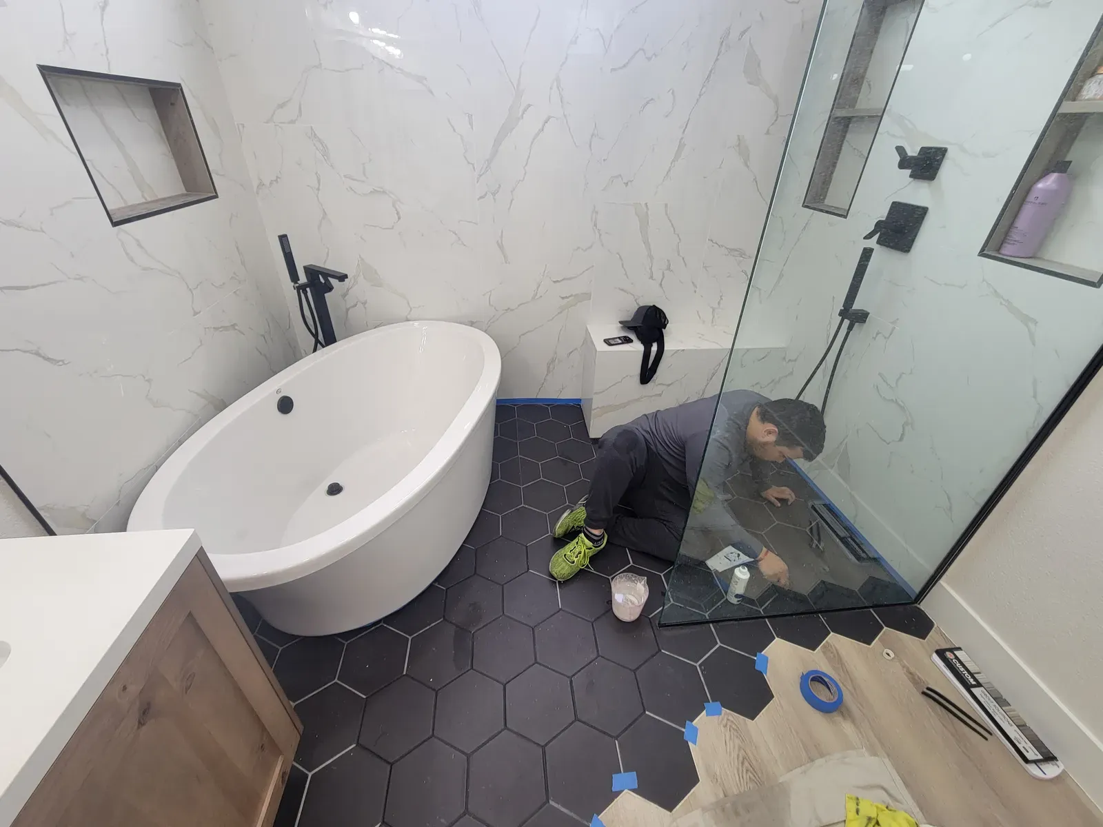 Technician sealing grout on a black hexagon tile bathroom floor beside a freestanding tub and glass shower in Las Vegas